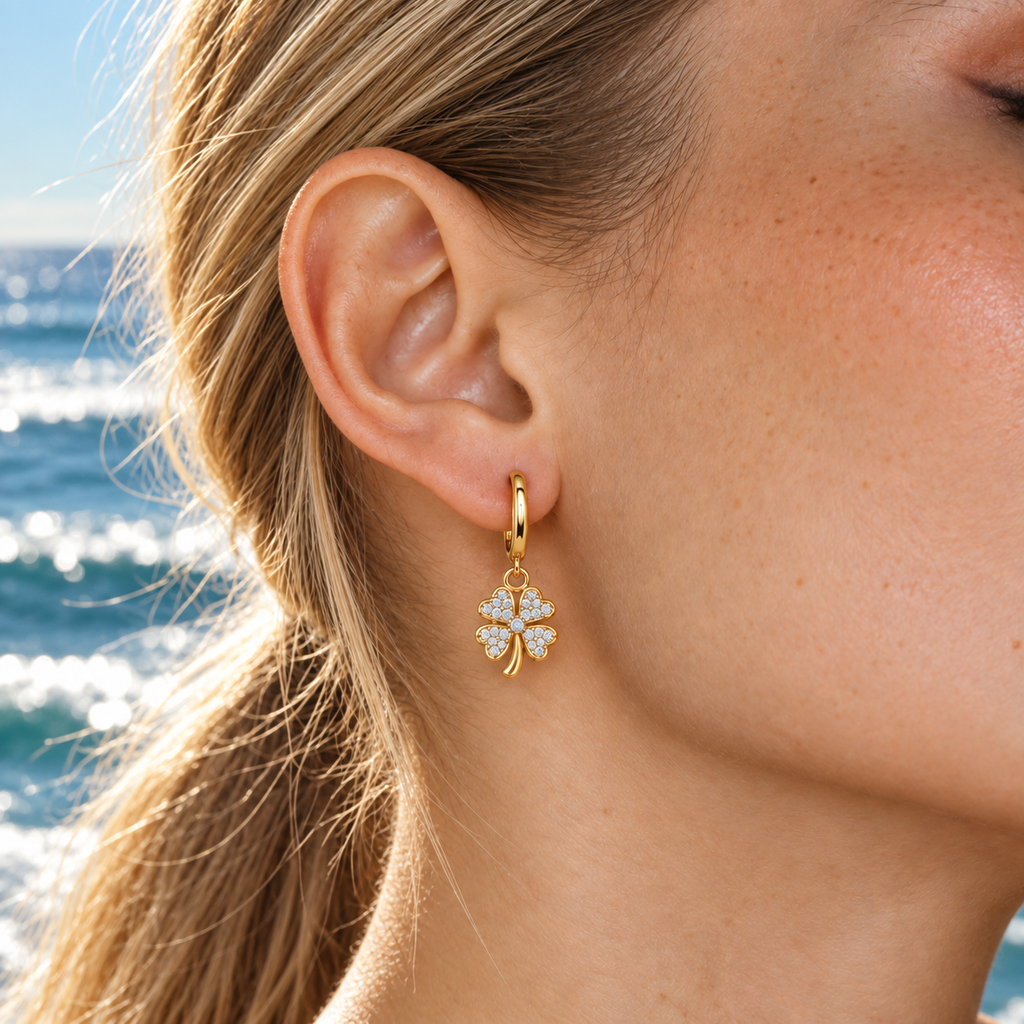 A close-up of a woman's ear wearing a gold huggie hoop earring with a dangling four-leaf clover charm set with clear crystals, with a blurred ocean background.