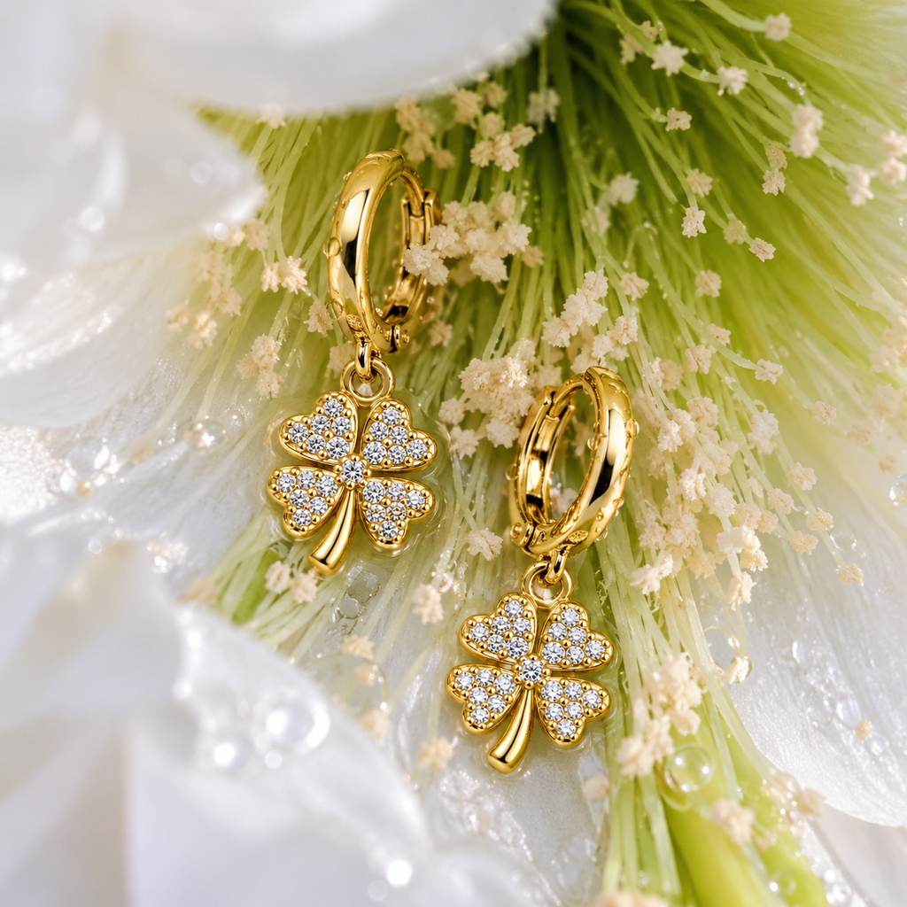 A pair of gold hoop earrings with dangling four-leaf clover charms paved with clear stones, displayed on a white flower with green stamens.