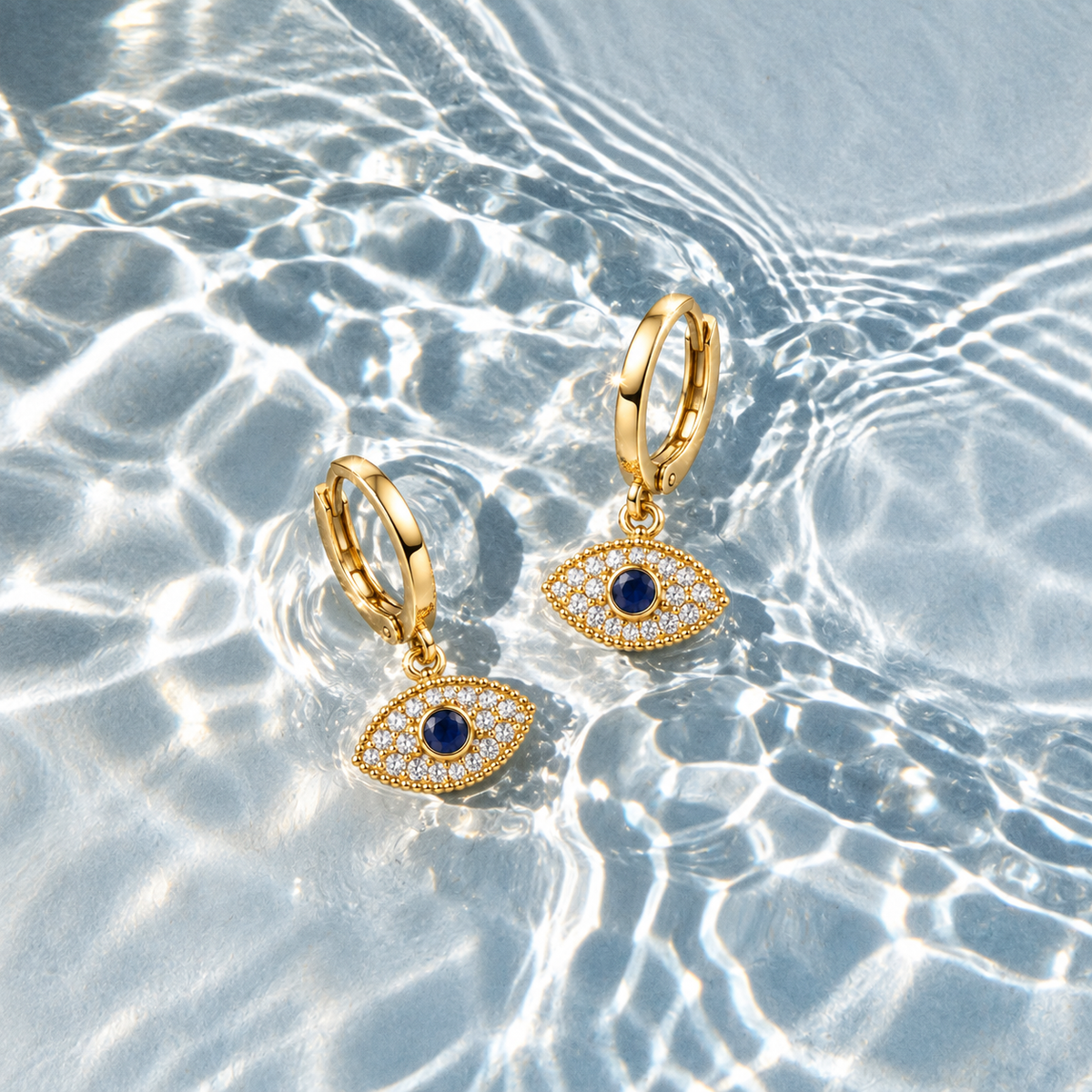 Gold huggie hoop earrings with evil eye charms featuring dark blue gemstones and clear pavé accents, displayed on a sunny, sunlit water surface with ripples and light reflections.