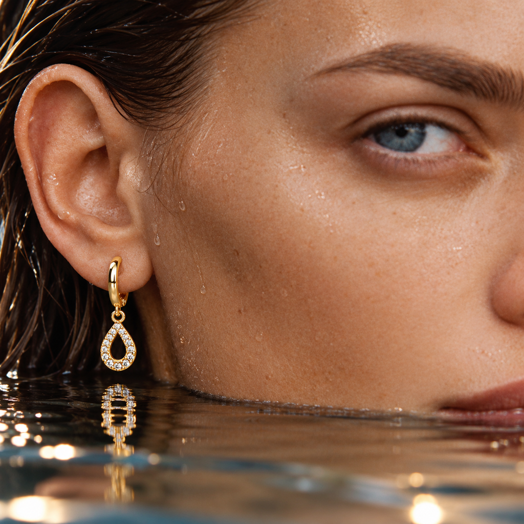 A close-up of a woman's ear wearing a gold huggie hoop earring with a teardrop-shaped crystal pavé setting, with water droplets on her face and waterline visible.
