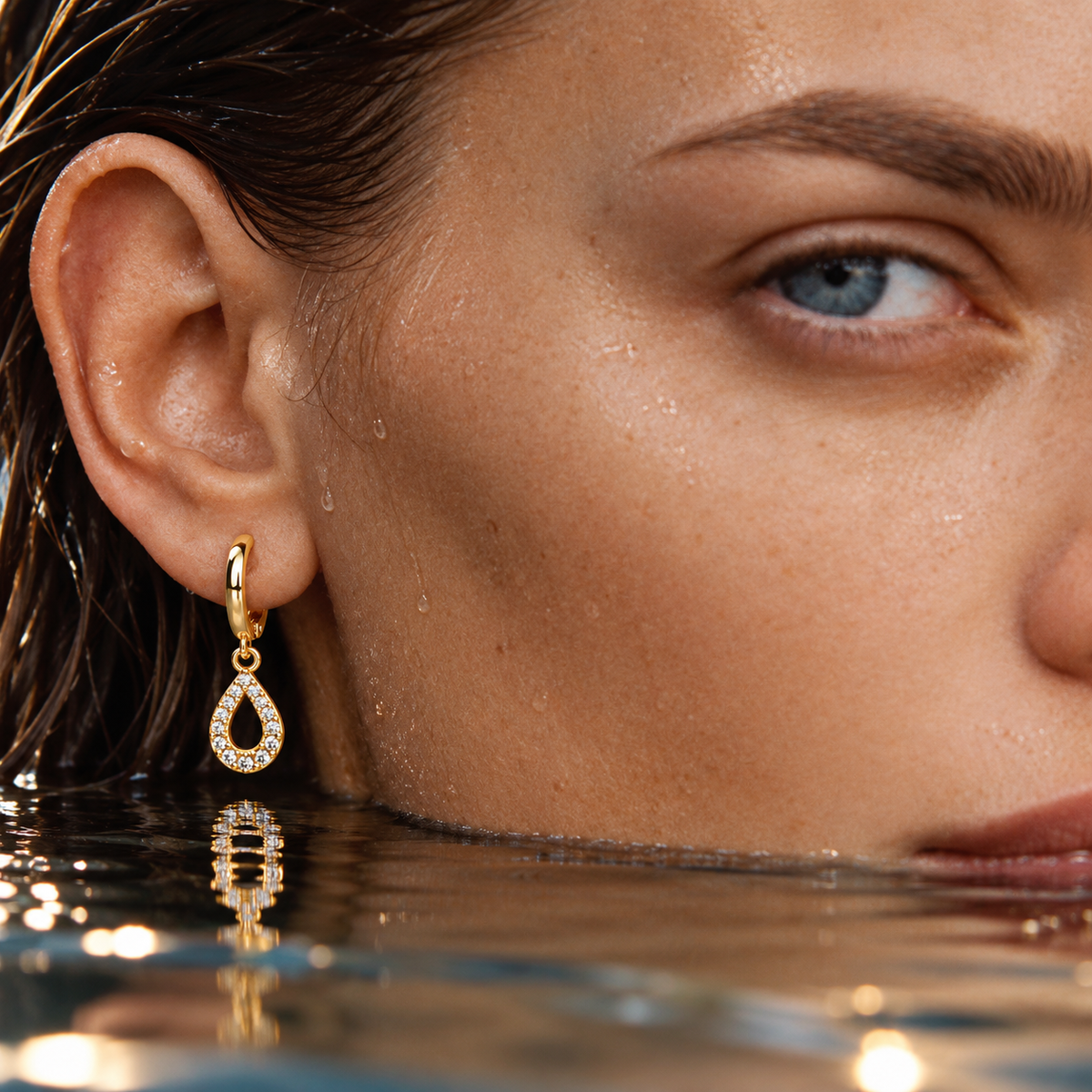 A close-up of a woman's ear wearing a gold huggie hoop earring with a teardrop-shaped crystal pavé setting, with water droplets on her face and waterline visible.