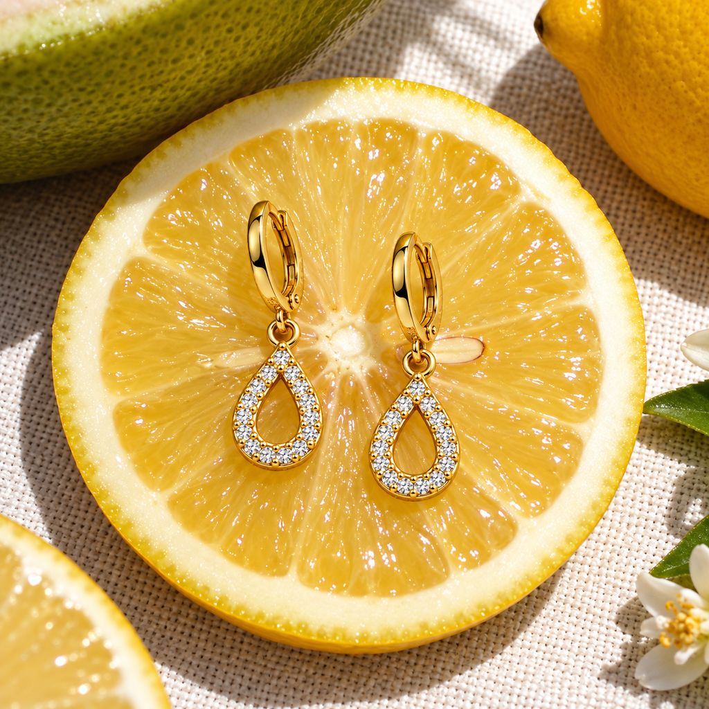 A pair of gold teardrop drop earrings with clear crystal pavé detailing, displayed on a fresh lemon slice with other lemons and a white flower on a textured beige fabric background.