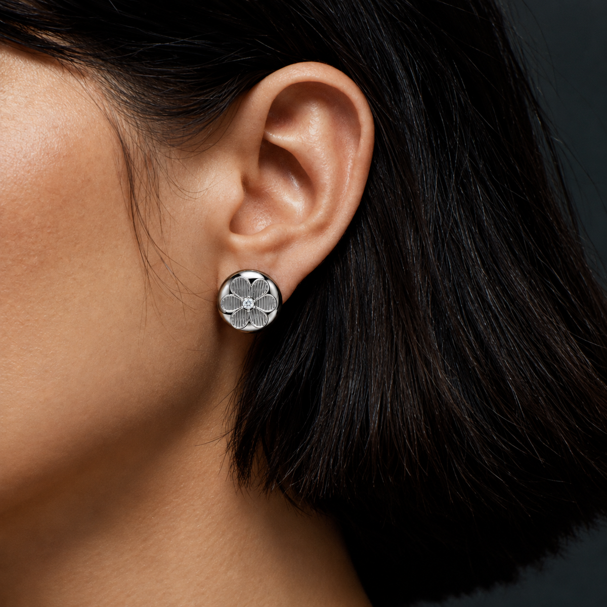 A close-up of a person's ear wearing a small round silver stud earring with a floral or clover-like cutout design, against dark hair and a dark background.