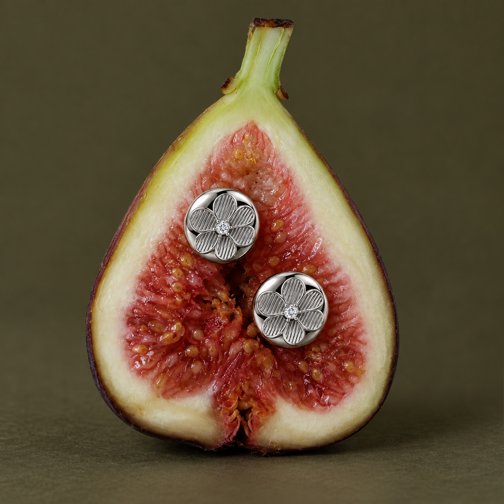 A close-up of a halved fig with two round silver flower-shaped stud earrings placed on the flesh. The background is a solid dark olive-brown tone.