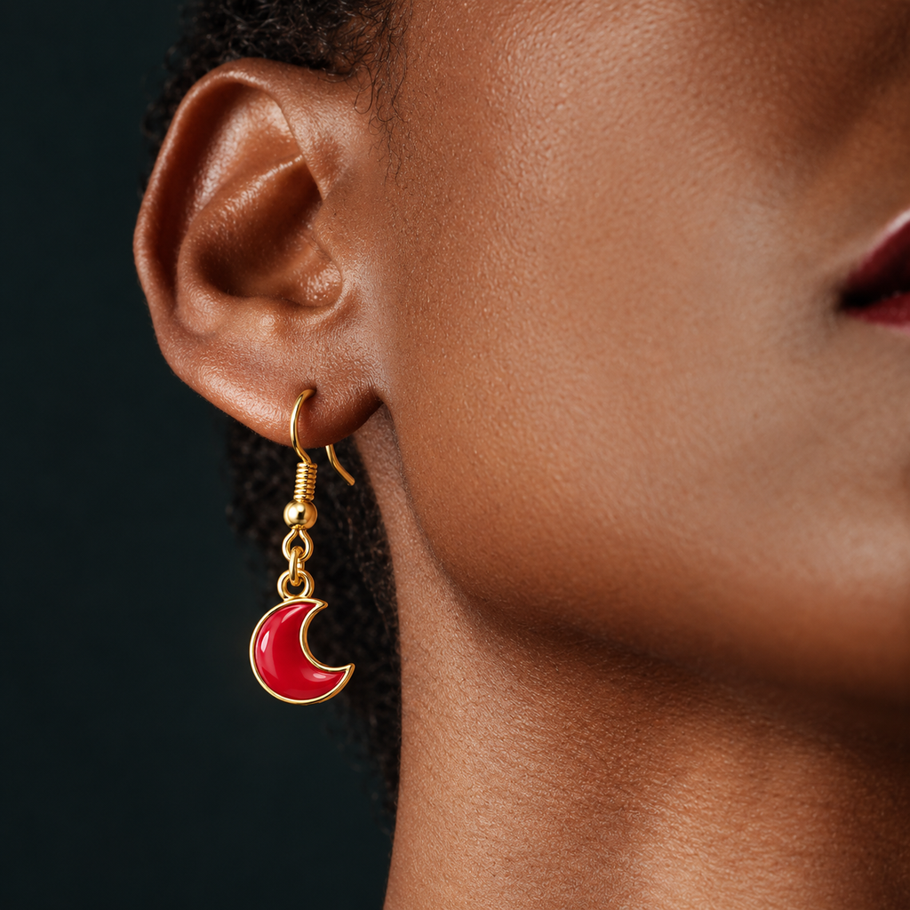 A close-up of a model's ear wearing a gold crescent moon dangle earring with a glossy red enamel center, set against a dark background.