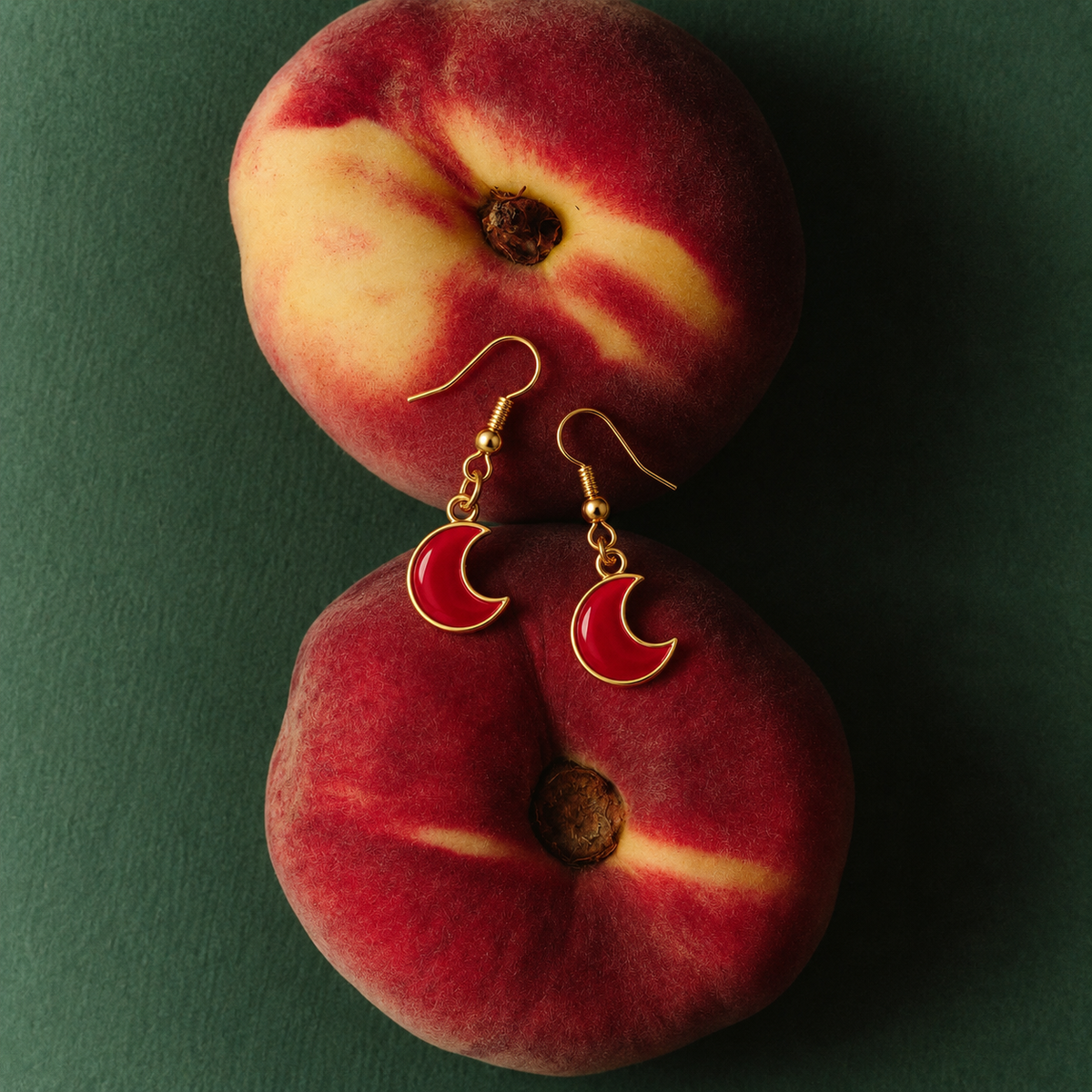 Gold crescent moon drop earrings with red enamel inlays, displayed on a dark green background with two whole peaches as props.