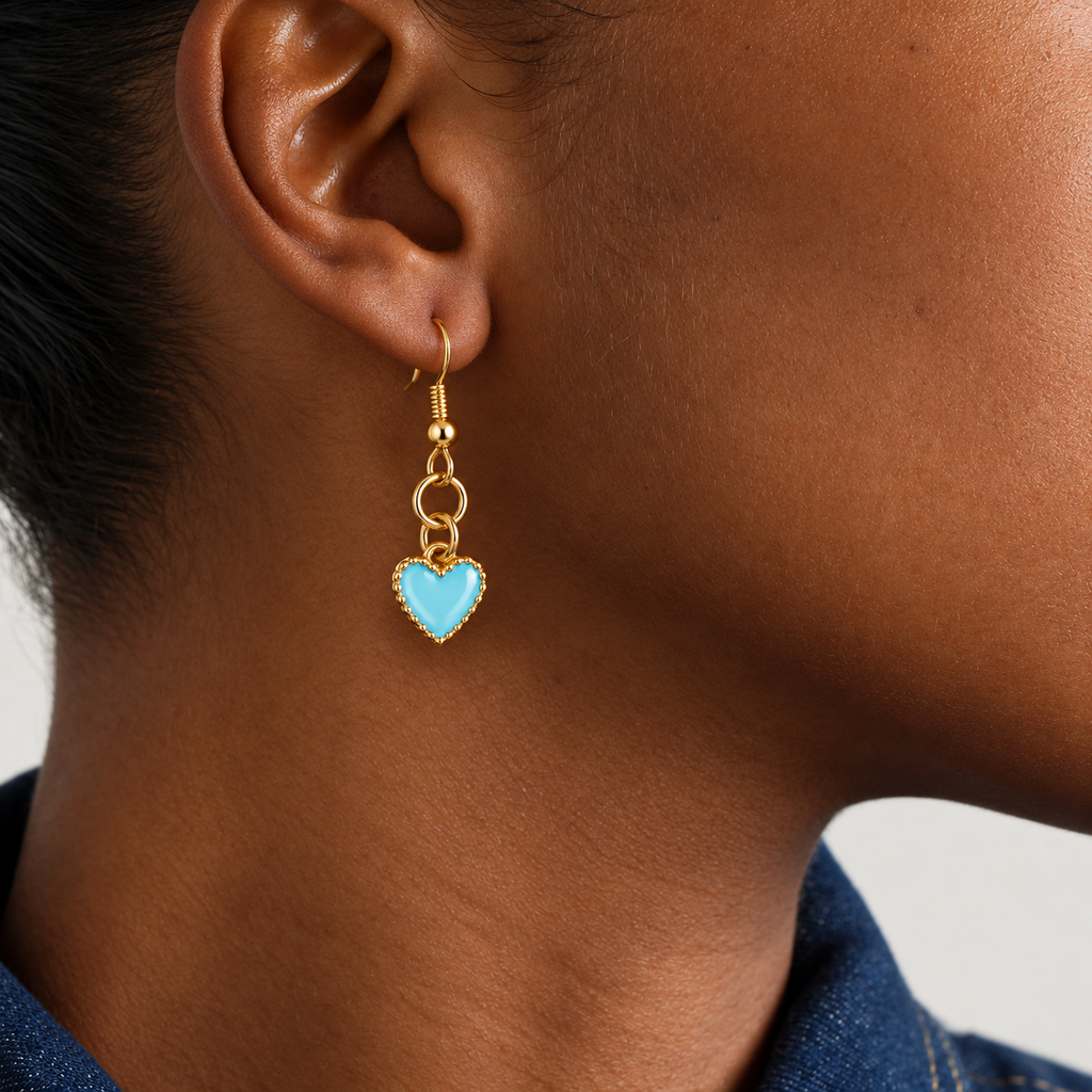 A close-up of a person's ear wearing a gold huggie hoop earring with a dangling heart charm. The heart charm has a light blue center with a delicate gold border.