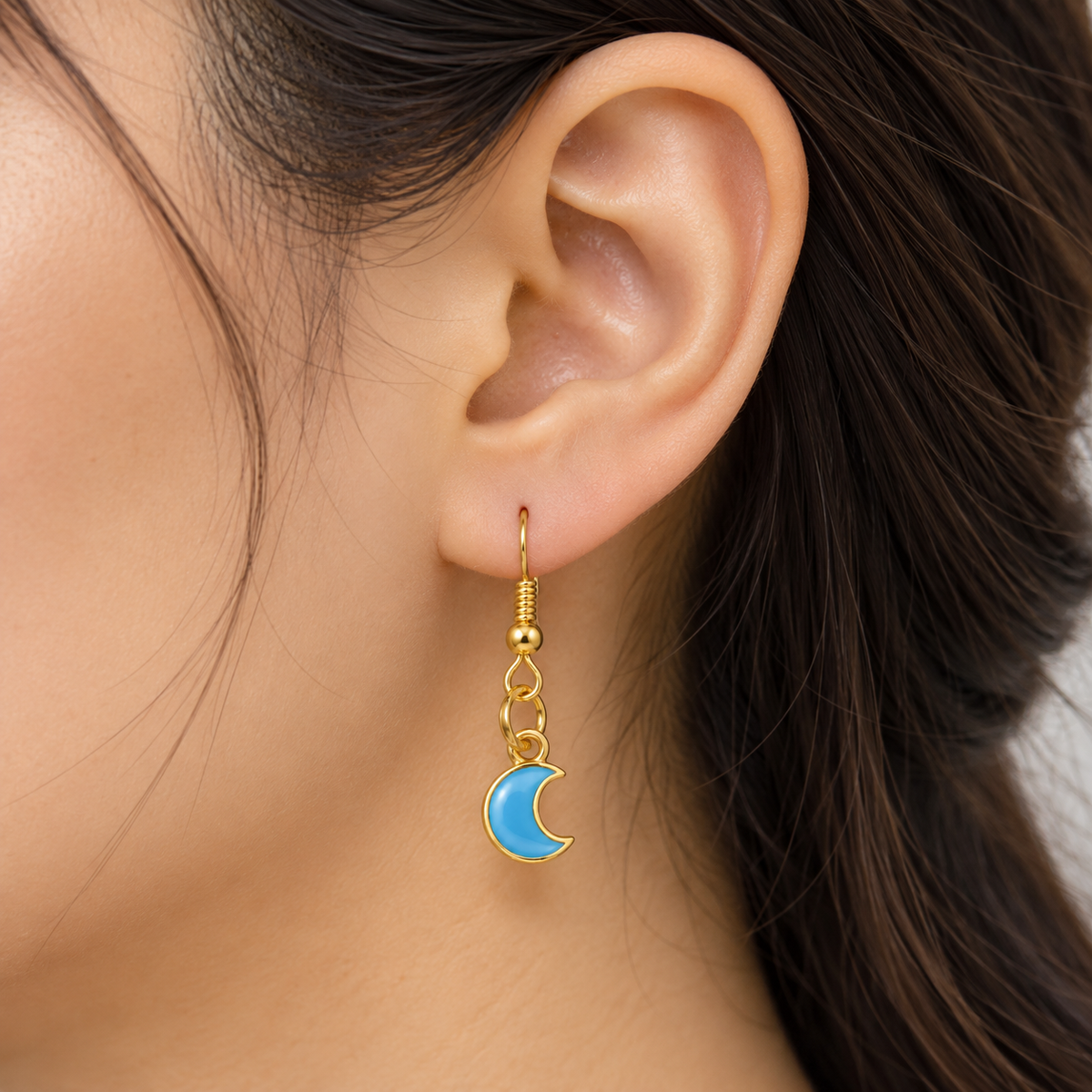 A close-up of a person's ear wearing a gold crescent moon dangle earring with light blue enamel.