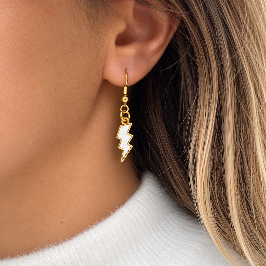 A close-up of a model's ear wearing a gold lightning bolt dangle earring with a white enamel center, paired with a white knit top.