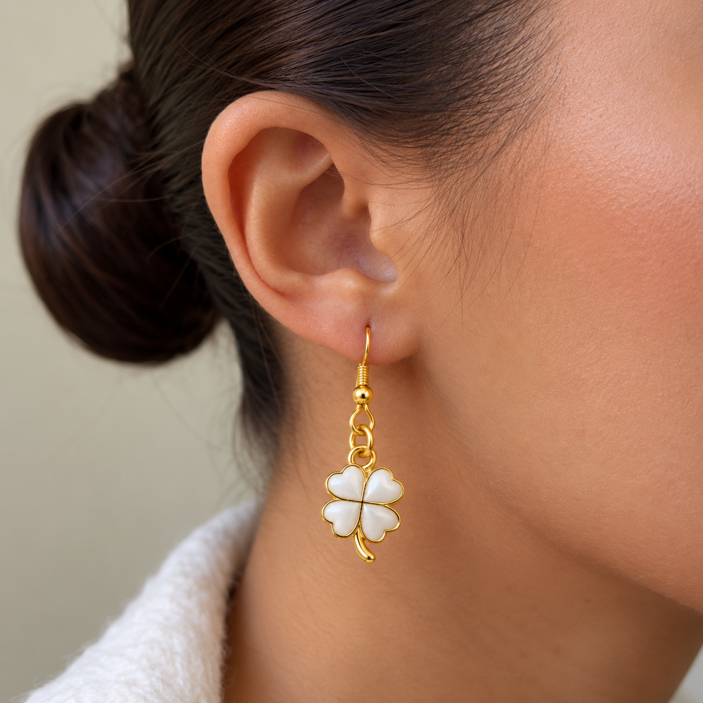 A close-up of a woman's ear wearing a dangling earring with a white four-leaf clover charm outlined in gold.