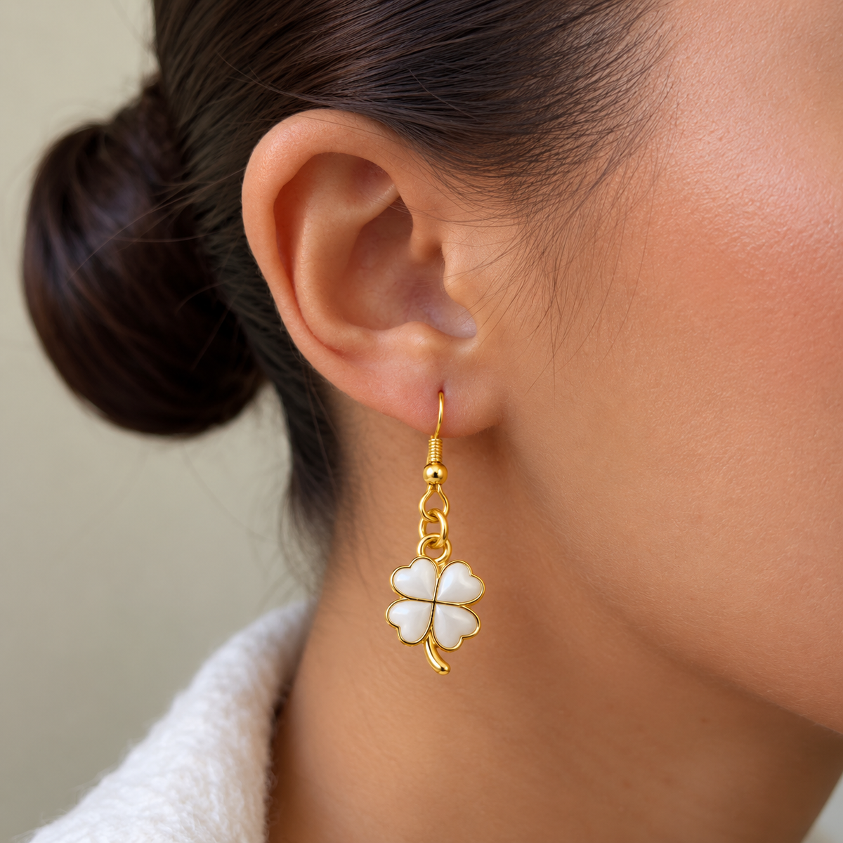 A close-up of a woman's ear wearing a dangling earring with a white four-leaf clover charm outlined in gold.