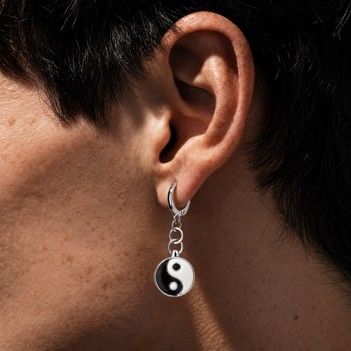 A close-up of a person's ear wearing a silver huggie hoop earring with a dangling charm shaped like a black-and-white yin-yang symbol.