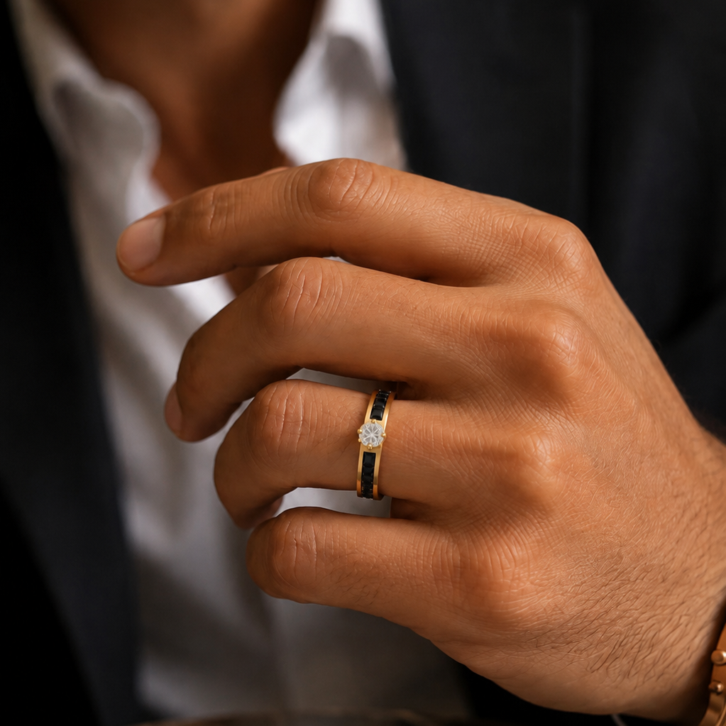 A close-up of a man's hand wearing a yellow gold ring with a black inlay and a central clear gemstone, styled with a white collared shirt and dark blazer.