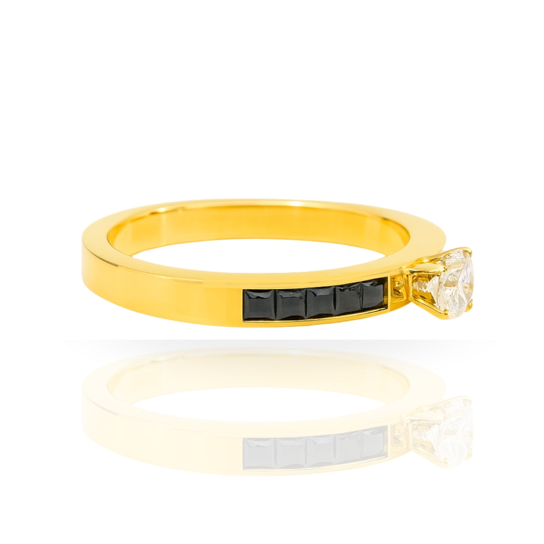 A yellow gold ring with a channel-set row of black stones and a single clear gemstone in a bezel setting, displayed on a clean white background with a soft reflection below.
