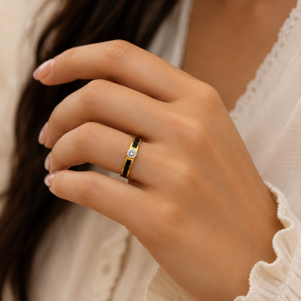 A close-up of a woman's hand wearing a yellow gold ring with a black inlay and a central clear gemstone, paired with a white lace-trim top.