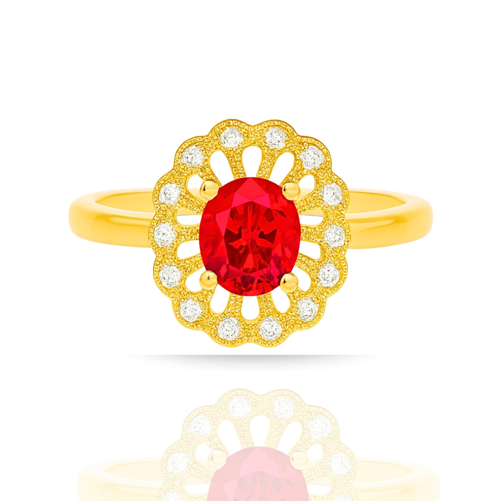 A yellow gold ring featuring an oval-cut red center stone surrounded by a scalloped halo of small clear stones, shown on a white background with a soft reflection below.