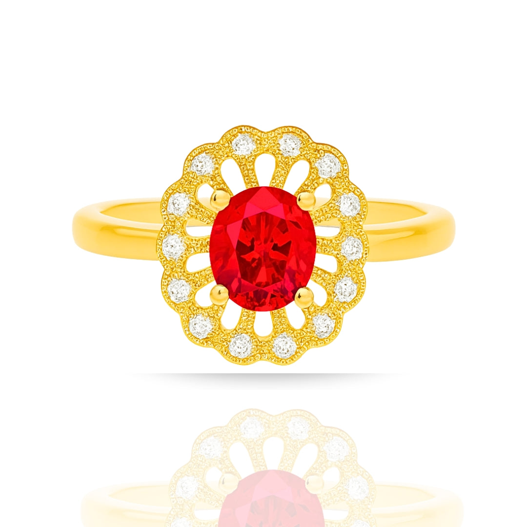 A yellow gold ring featuring an oval-cut red center stone surrounded by a scalloped halo of small clear stones, shown on a white background with a soft reflection below.