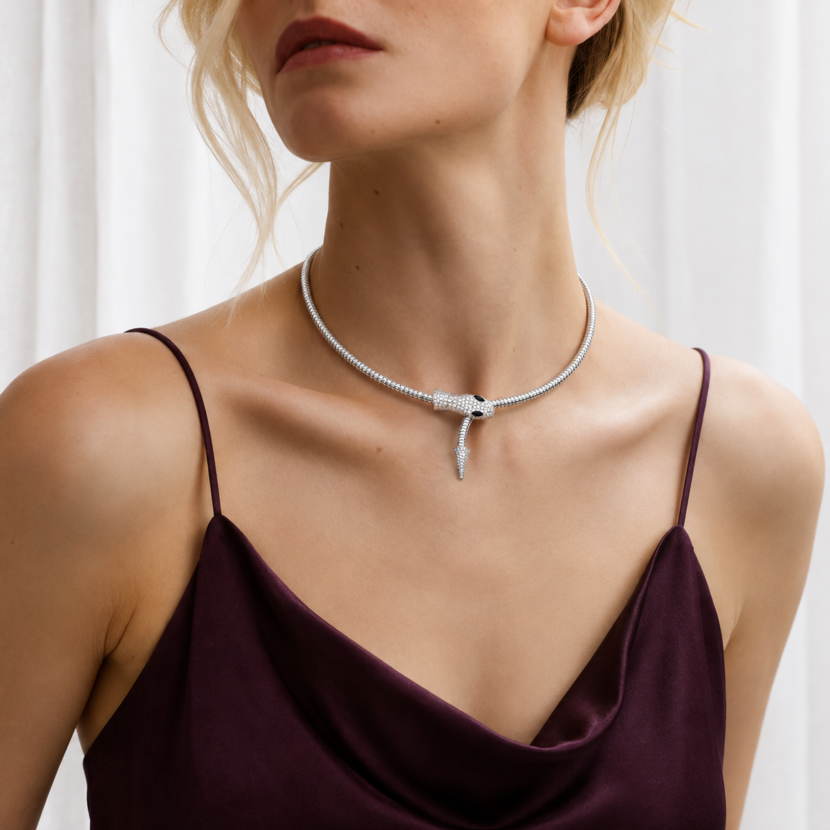 A close-up of a model wearing a silver snake choker necklace with a diamond-encrusted head, paired with a dark plum satin camisole top.