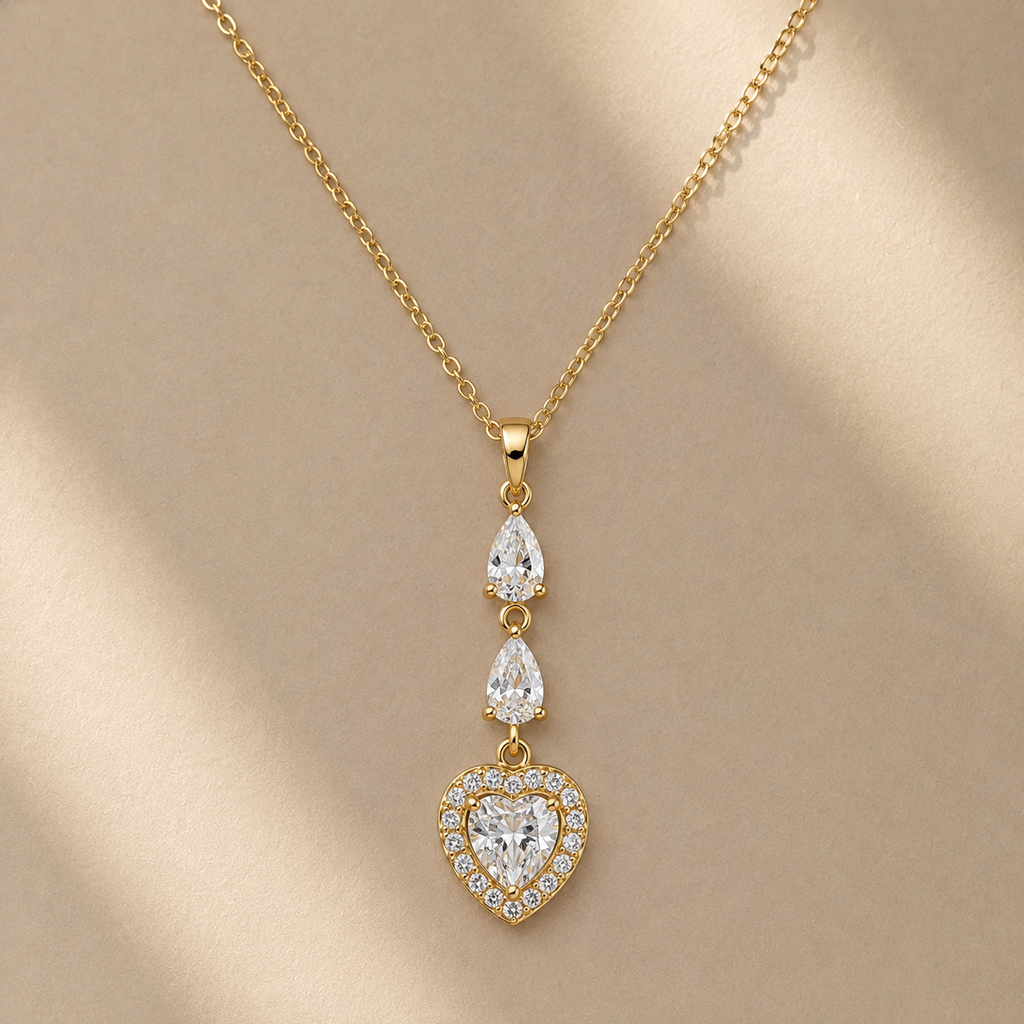 Gold-tone dangle necklace with a heart-shaped pendant and two teardrop-shaped clear stones, displayed on a beige background with soft diagonal shadows.