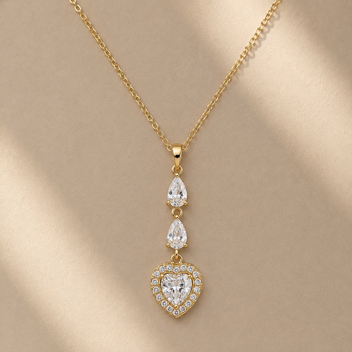 Gold-tone dangle necklace with a heart-shaped pendant and two teardrop-shaped clear stones, displayed on a beige background with soft diagonal shadows.