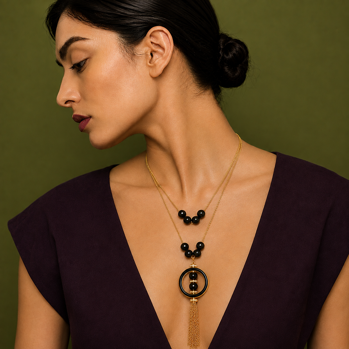 A woman in profile wearing a gold-toned double-strand necklace with black beads and a circular pendant, paired with a dark purple V-neck top against an olive green background.