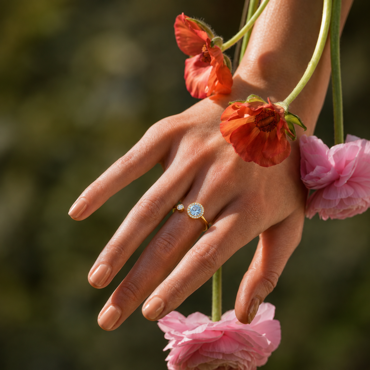 A close-up of a hand wearing a delicate gold ring with a round clear gemstone and a small white accent, posed among pink and orange flowers against a soft green blurred background.