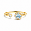 A gold open-shank ring featuring a large round light blue center stone surrounded by a halo of small clear stones, with a smaller matching round stone on the opposite side of the band, shown on a white background.
