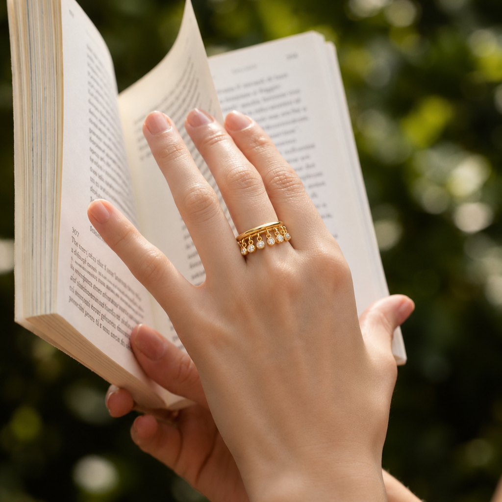 A close-up lifestyle shot of a person's hand holding an open book, wearing a delicate gold band ring adorned with small white pearls on the ring finger. The background is a soft, out-of-focus green foliage.