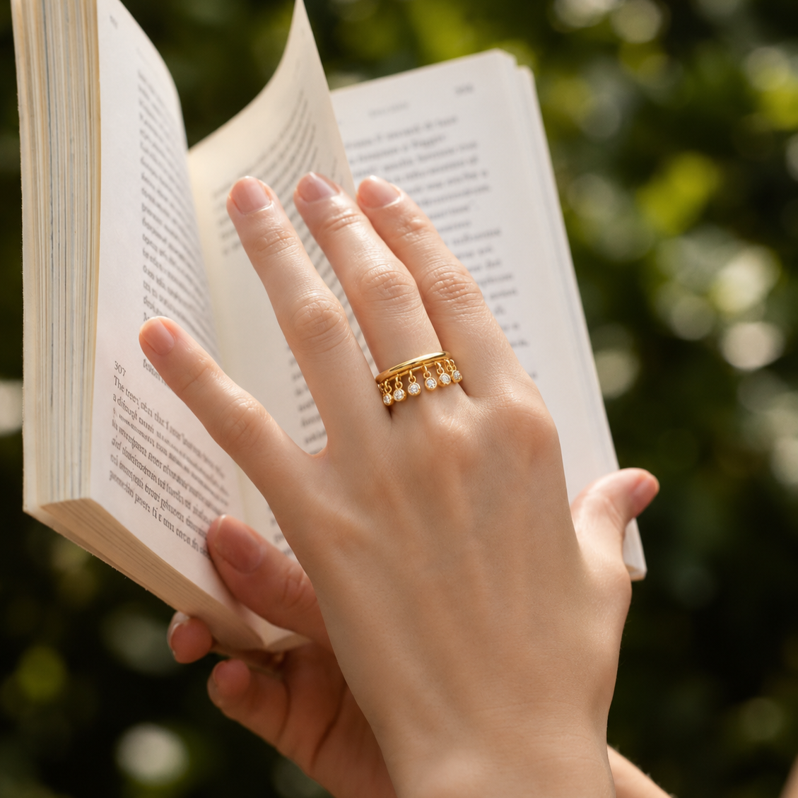 A close-up lifestyle shot of a person's hand holding an open book, wearing a delicate gold band ring adorned with small white pearls on the ring finger. The background is a soft, out-of-focus green foliage.