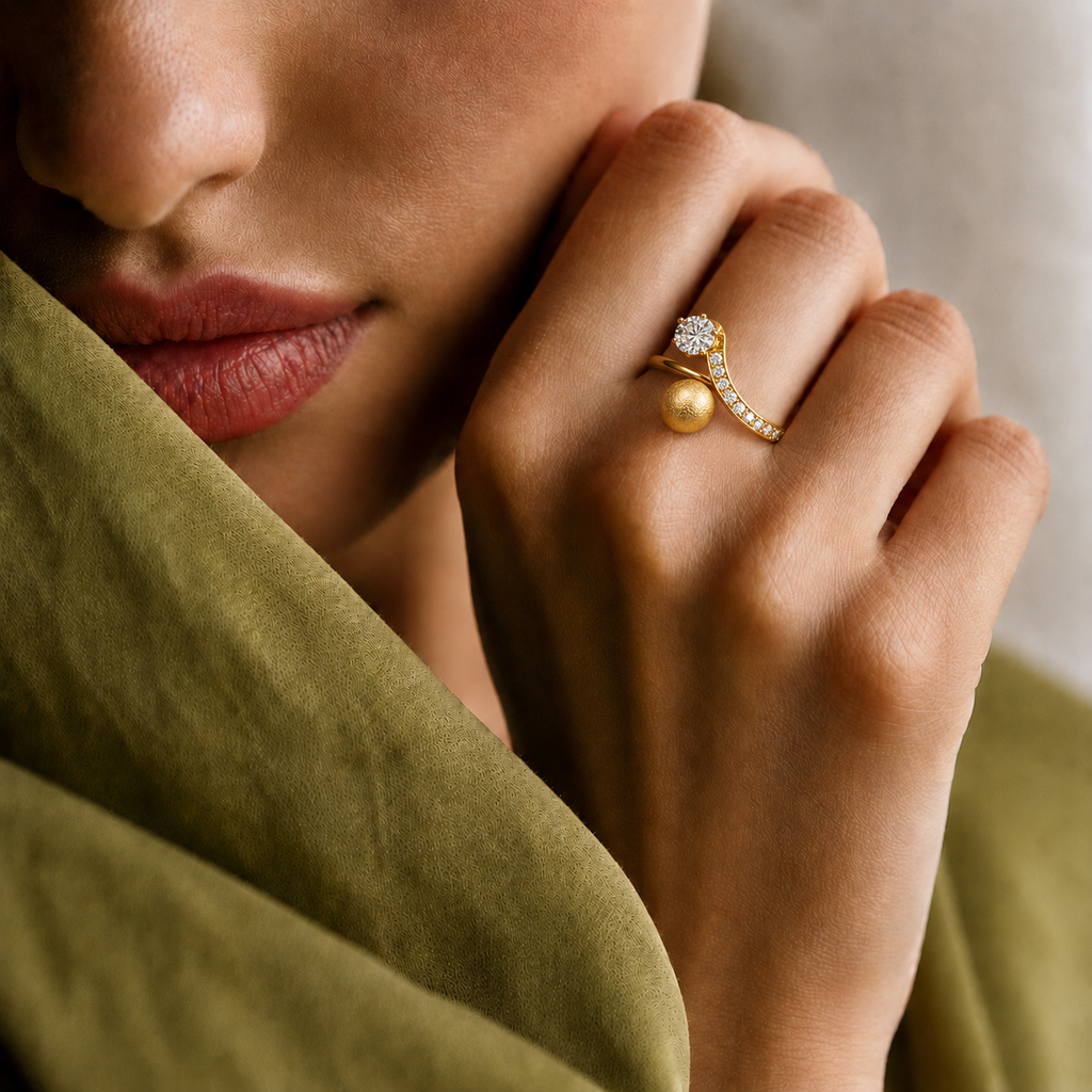 A close-up lifestyle shot of a model wearing a gold ring with a round pearl and a small diamond cluster, styled with an olive green fabric drape.