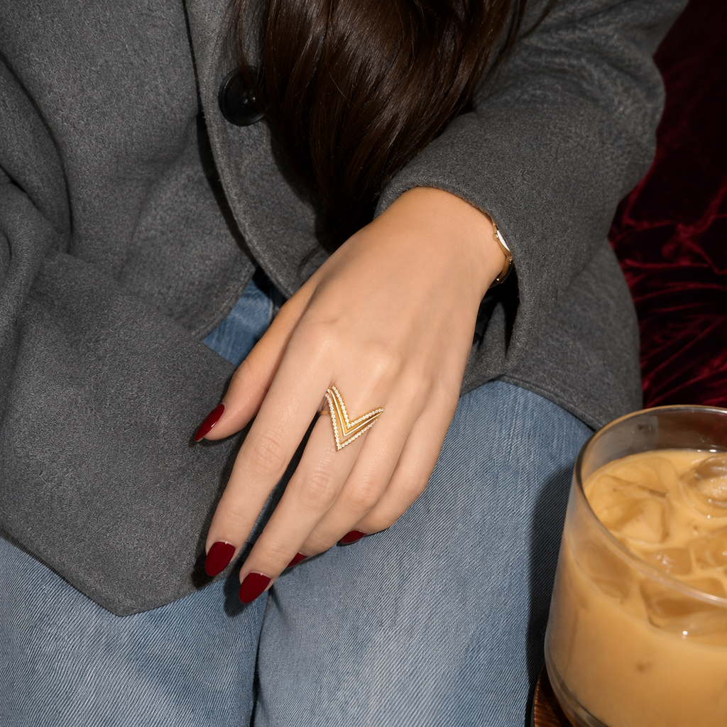 A close-up lifestyle shot of a person wearing a gold chevron ring on their hand, dressed in a gray blazer and blue jeans, with an iced coffee in the foreground.