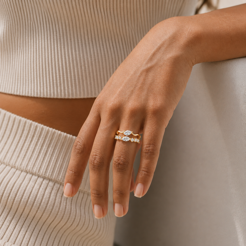 A close-up of a person's hand wearing a delicate gold ring with a marquise-cut center stone and small side stones, styled with a ribbed beige top and skirt.