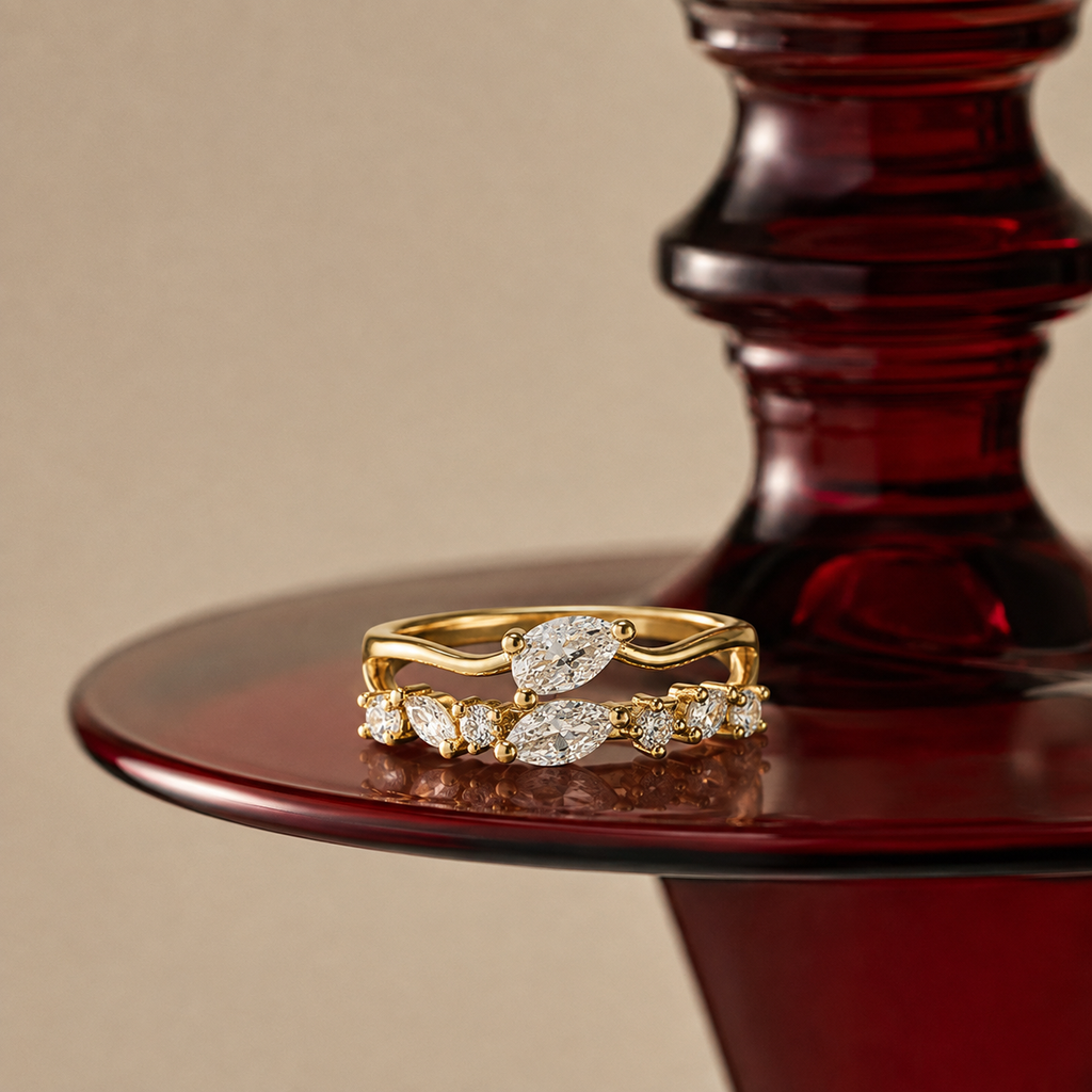A pair of yellow gold rings with marquise-cut and round diamonds, displayed on a dark red glass pedestal against a warm beige background.