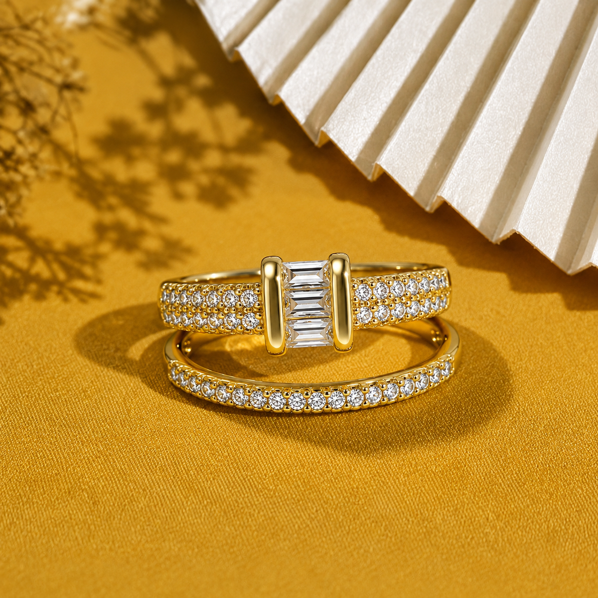 A pair of gold rings with pavé-set clear stones, featuring a central baguette-cut diamond on the top ring and a smaller band ring, displayed on a textured gold surface with a white fan in the background.