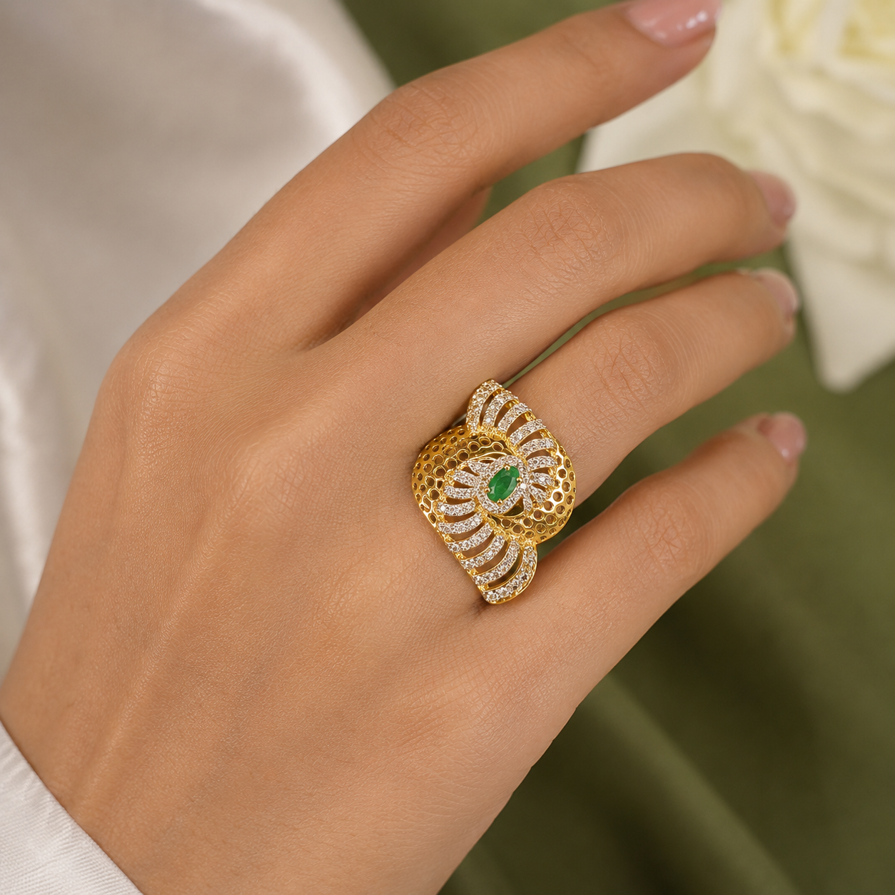 A close-up of a hand wearing a large gold ring with a central green oval stone and pavé-set clear stones around it, against a soft white and olive green background.