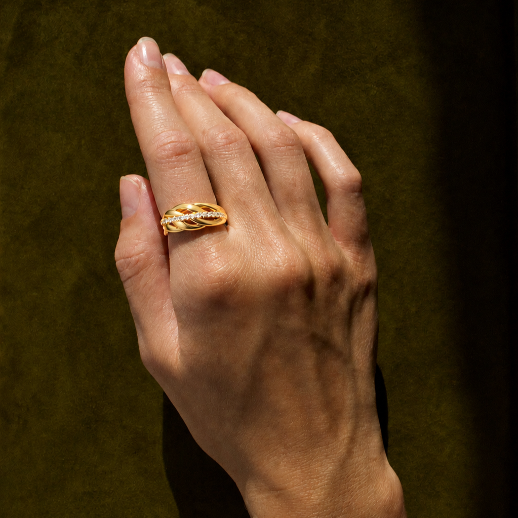 A close-up of a hand wearing a gold ring with a textured, leaf-like design and small clear stones, set against a dark olive green background.