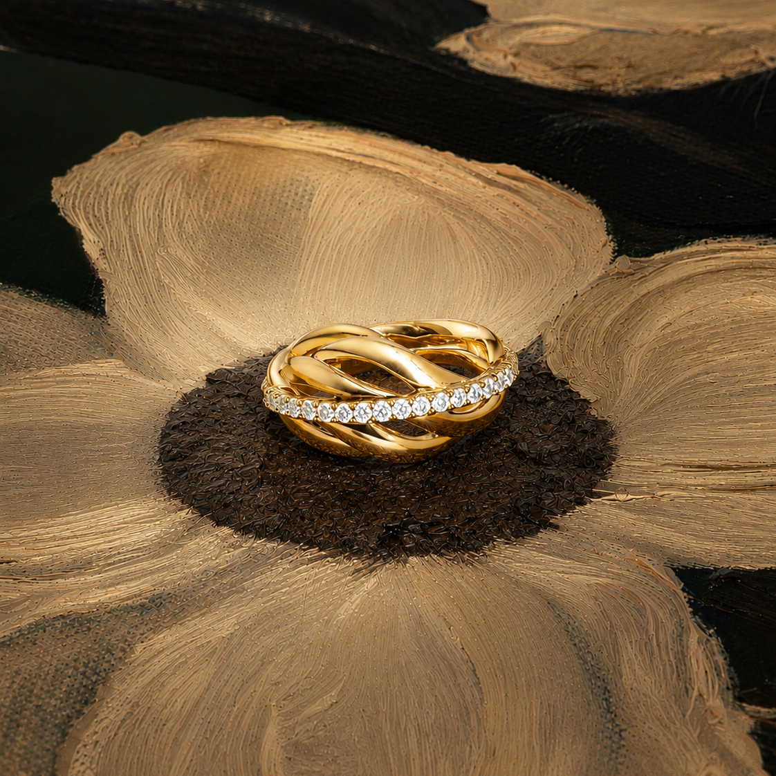 A gold ring with a twisted knot design and a row of small clear stones on one side, displayed on a textured gold flower against a dark background.